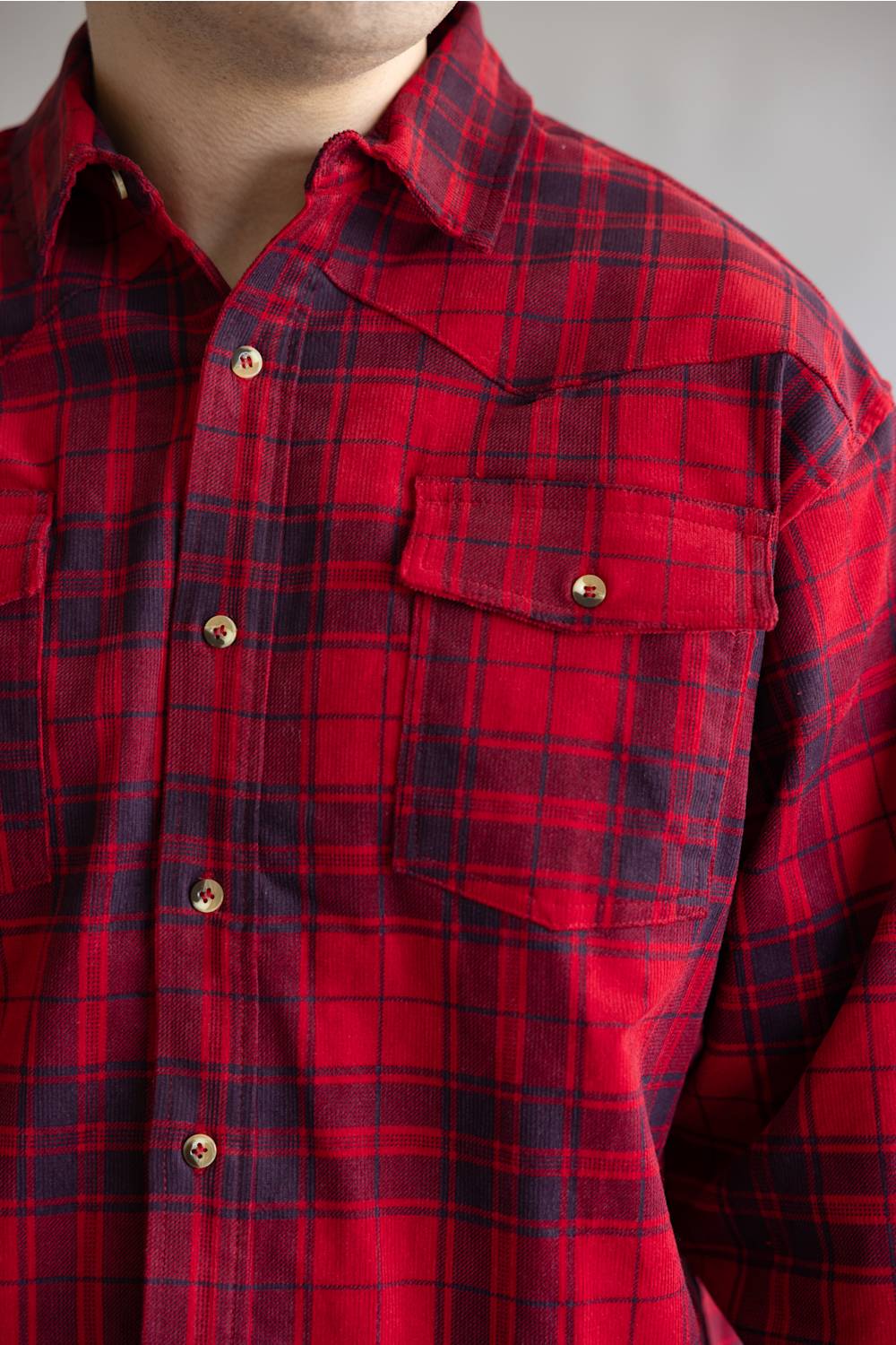 A close-up of a red and black plaid button-down shirt worn by a model, showing the collar, chest pocket with a snap button, and fabric texture.