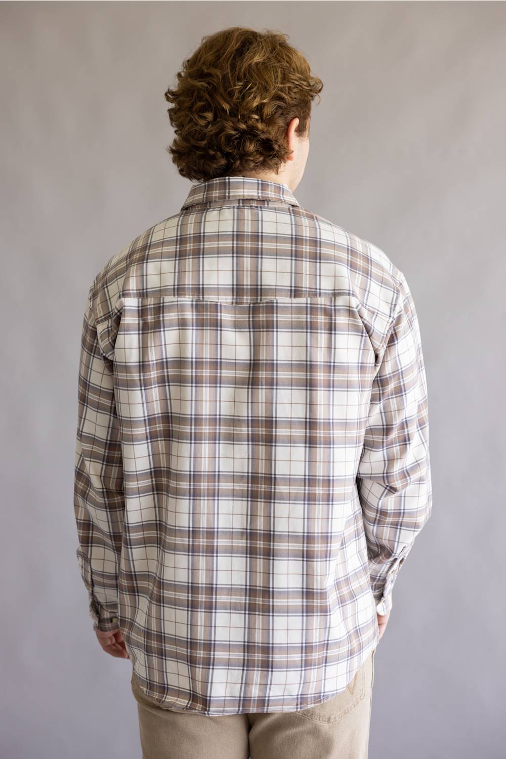 A person viewed from behind wearing a long-sleeve plaid button-up shirt in off-white with brown and gray lines, paired with khaki pants, against a plain gray studio background.