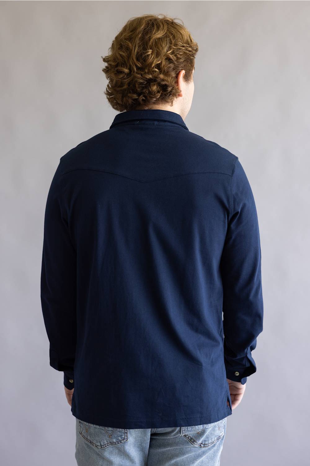 A man viewed from behind wearing a dark navy blue long-sleeve button-up shirt and light blue jeans, standing against a neutral gray background.