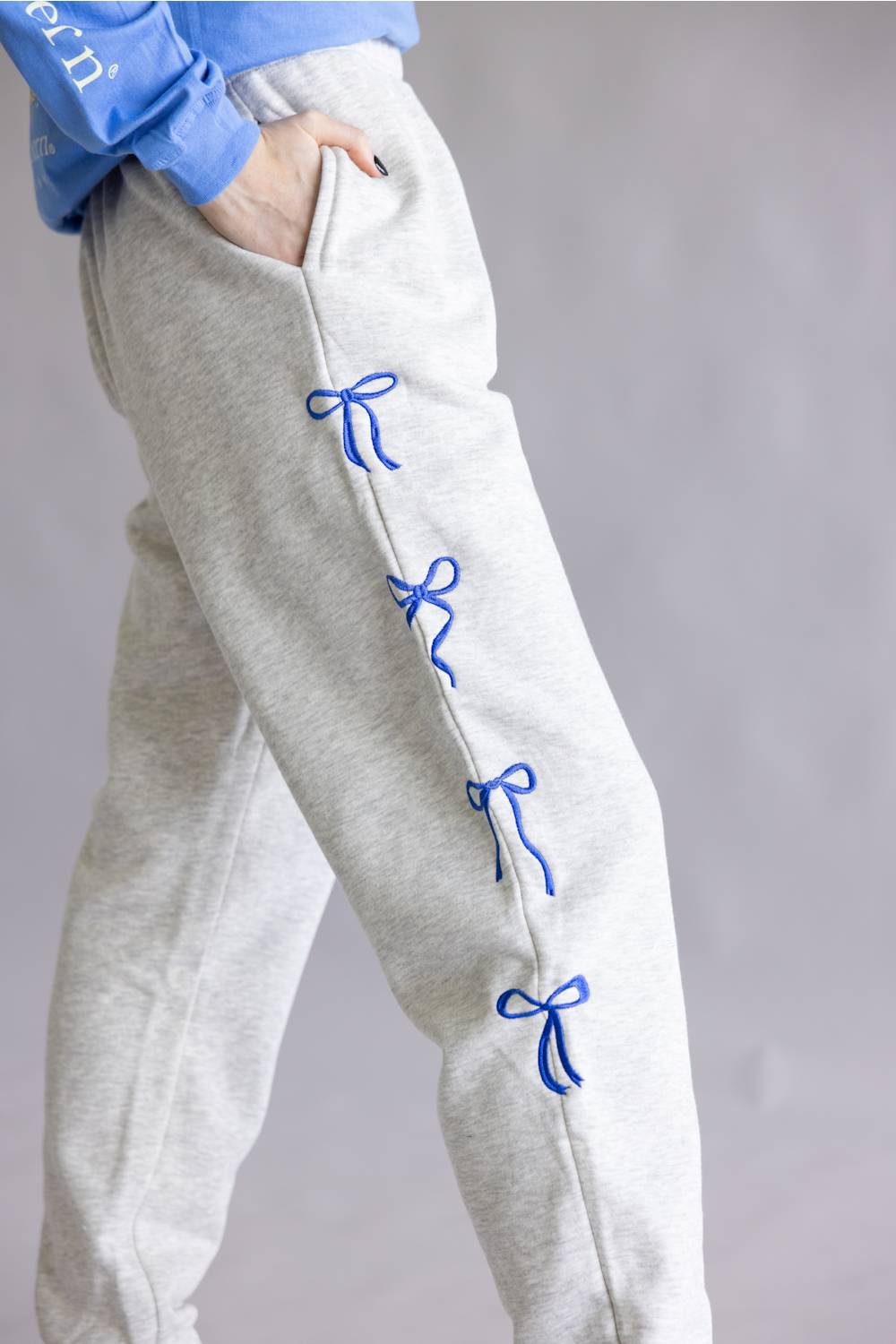 Close-up of a person wearing light heather gray sweatpants with blue embroidered bow details down the side, paired with a blue top against a soft gray background.