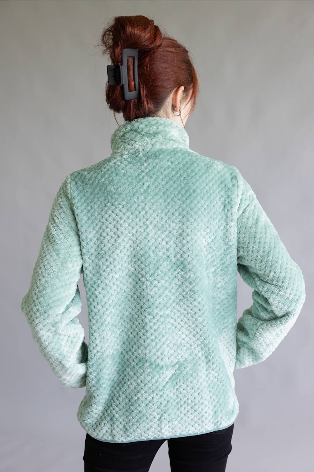 A person shown from the back wearing a textured mint green fleece pullover jacket, paired with black pants, against a plain gray studio background.