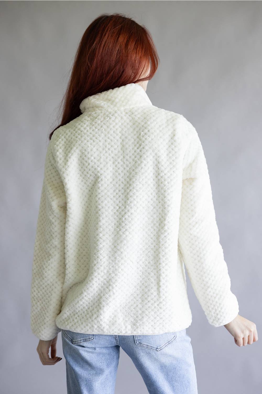 A person wearing a cream-colored textured fleece pullover jacket with a half-zip collar, a chest pocket, and a brown rectangular patch on the upper chest, paired with blue jeans against a neutral gray background.