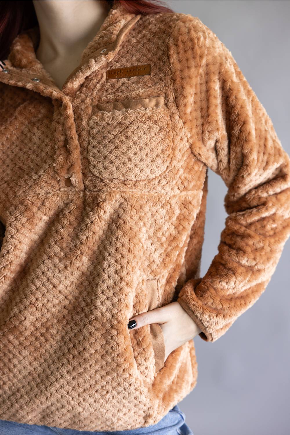 A close-up of a person wearing a textured, waffle-knit fleece pullover in a warm camel brown color, paired with blue jeans against a neutral gray background.