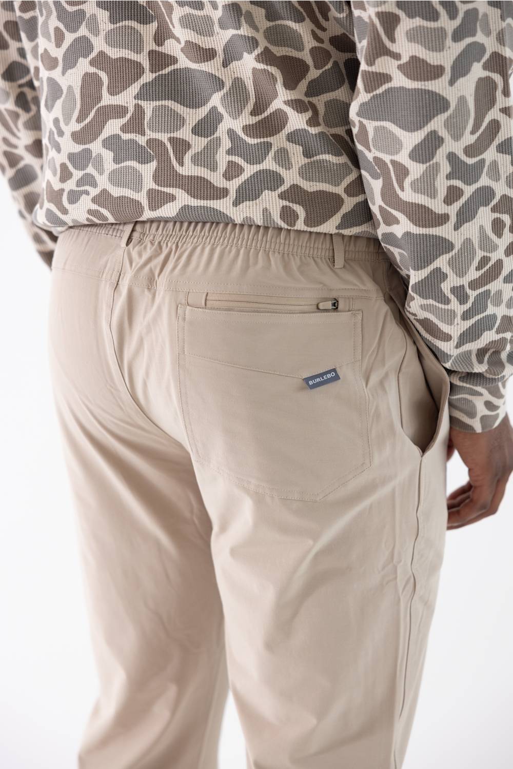 Close-up back view of a person wearing beige drawstring pants with a small blue brand tag on the back pocket, paired with a beige and gray patterned long-sleeve top against a white studio background.