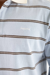 Close-up of a light blue striped polo shirt with thin brown horizontal stripes and a small white embroidered script logo on the chest.