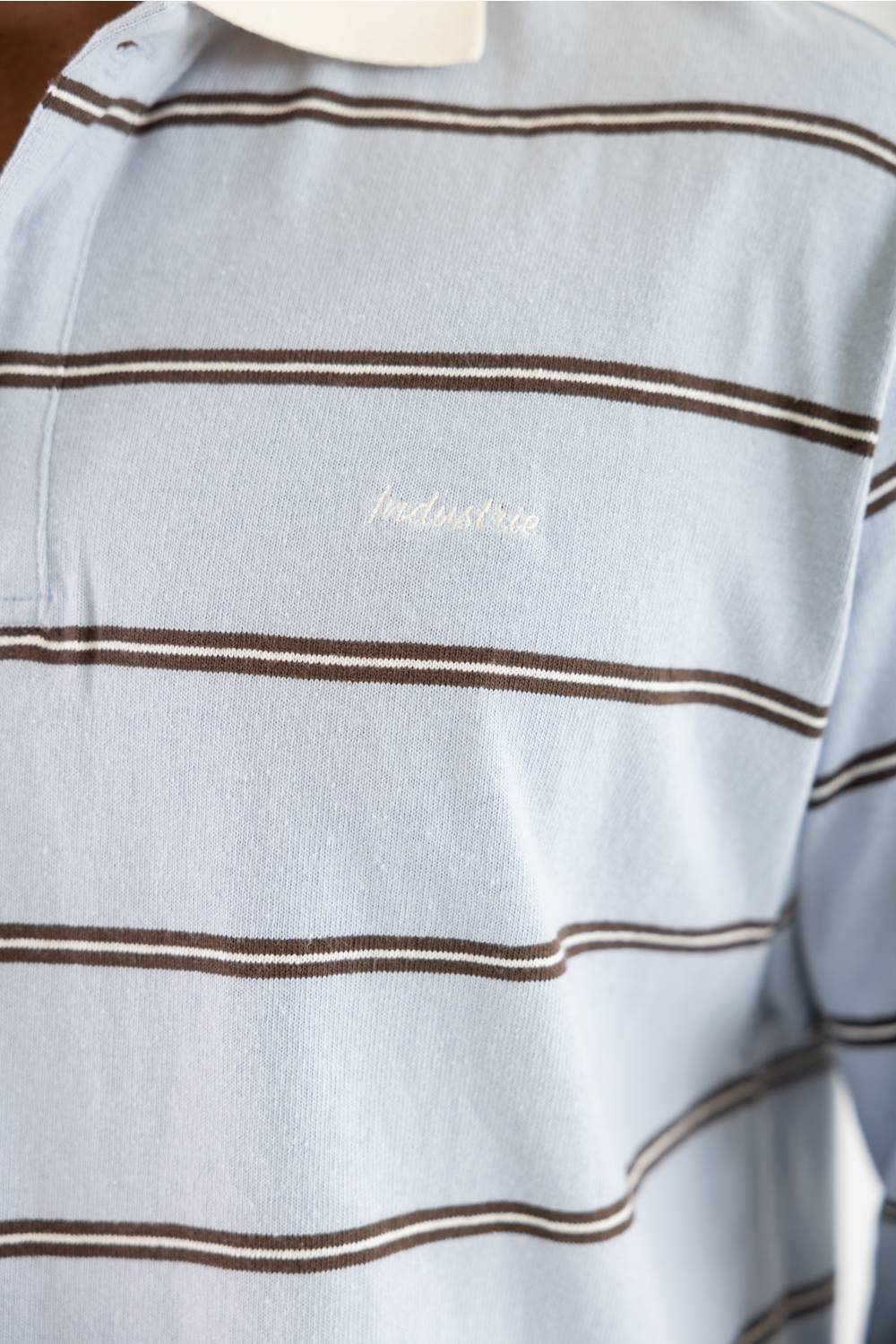 Close-up of a light blue striped polo shirt with thin brown horizontal stripes and a small white embroidered script logo on the chest.