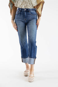 A model wearing a pair of medium-wash blue cropped jeans with rolled cuffs, paired with a patterned top and beige flat shoes, photographed against a plain white studio background.