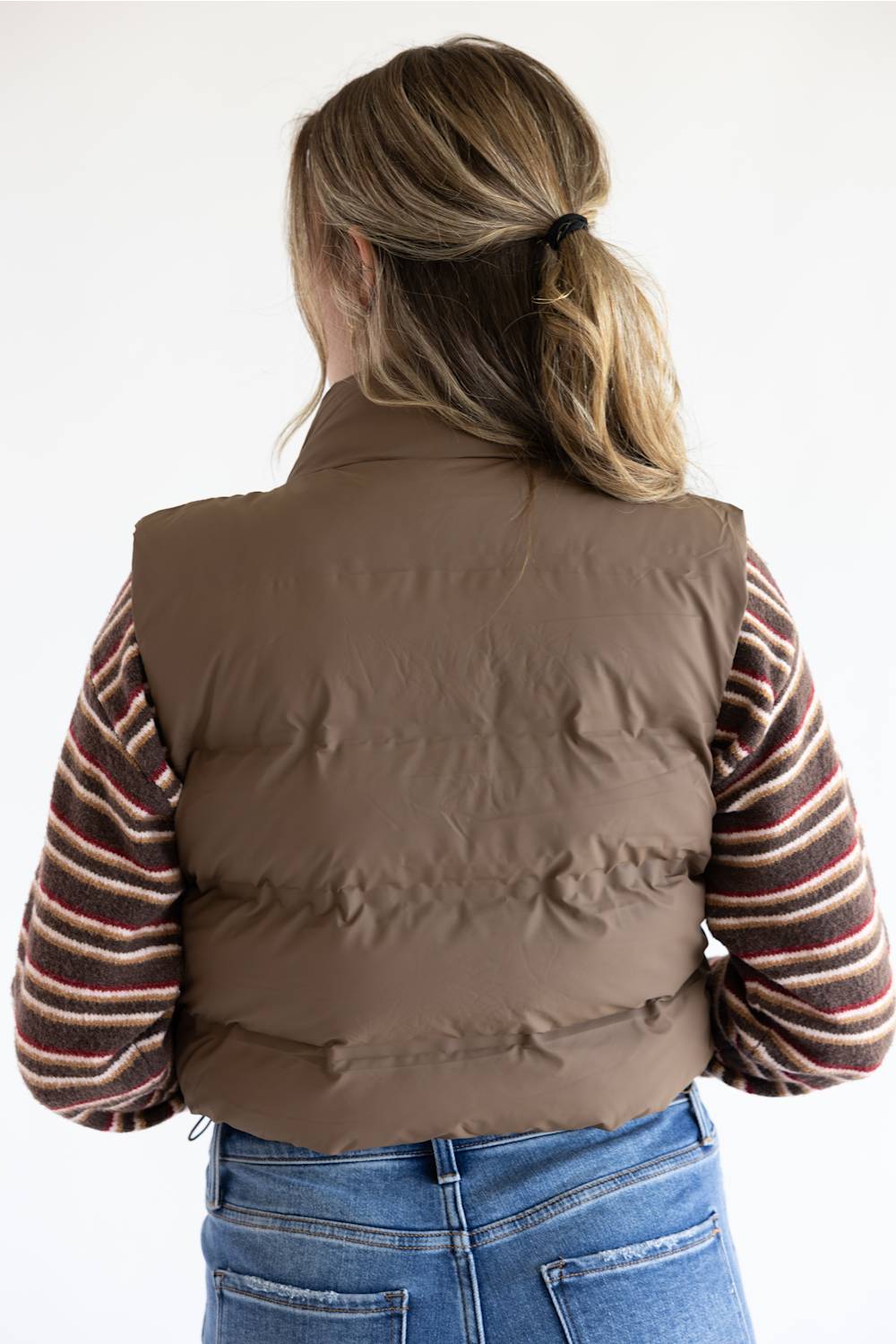 A woman viewed from behind wearing a brown puffer vest over a striped long-sleeve shirt and blue jeans against a plain white background.