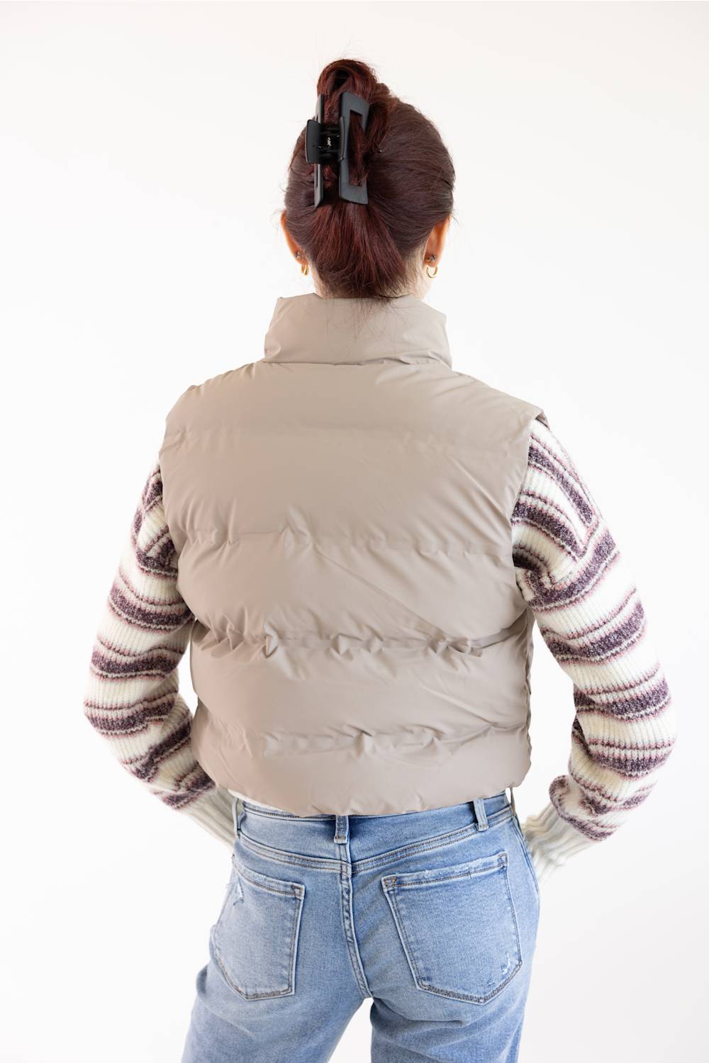 A person shown from the back wearing a light beige puffer vest over a purple and cream striped knit sweater, paired with light blue jeans, against a plain white background.