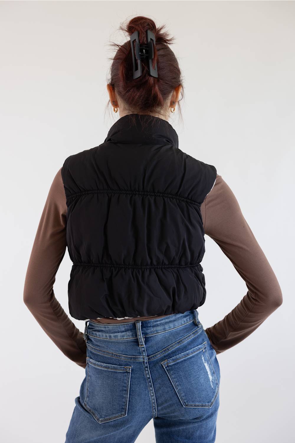 A woman shown from behind wearing a black quilted sleeveless puffer vest over a brown long-sleeve top and blue jeans, against a plain light background.