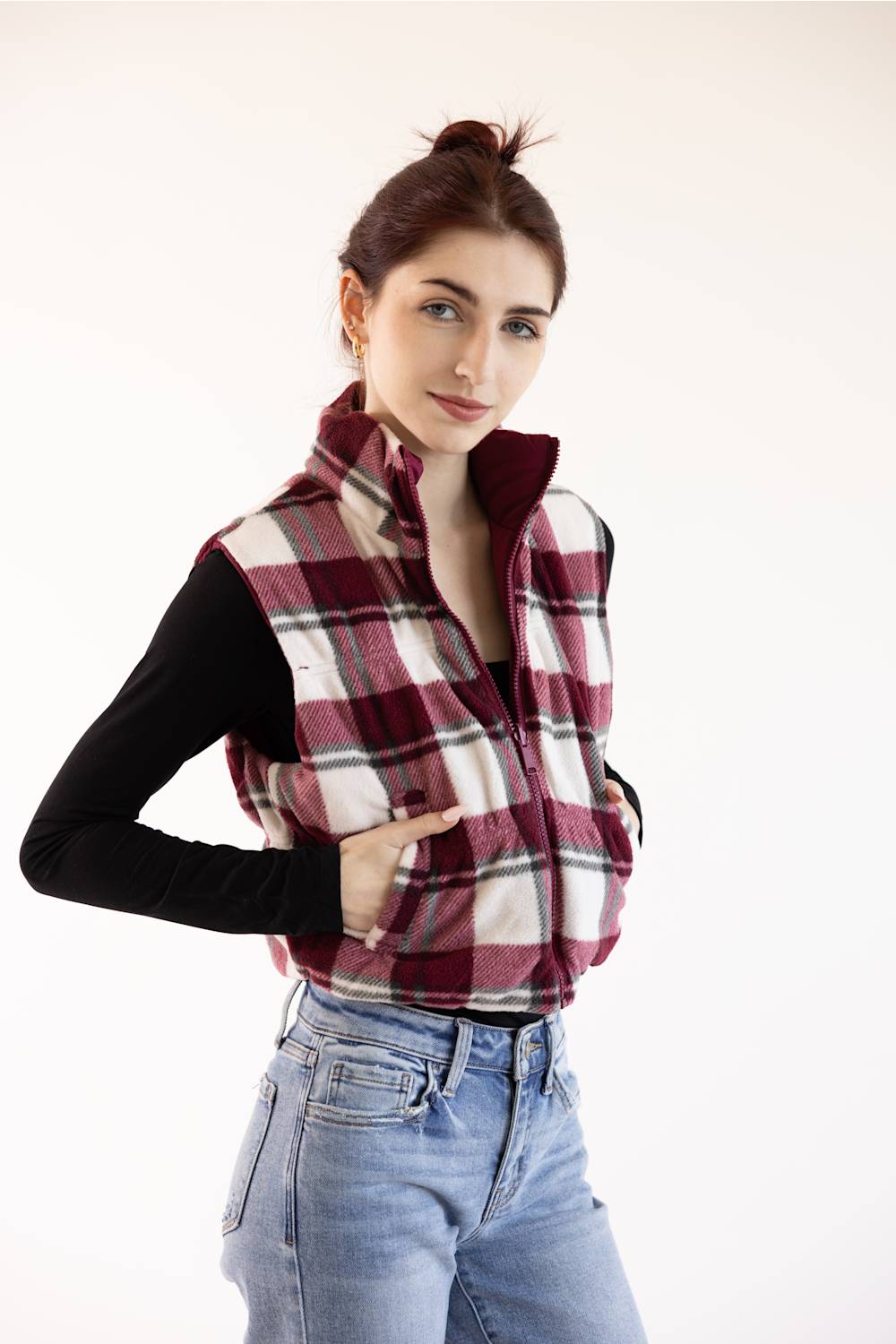 A woman wearing a burgundy, white, and black plaid zip-up fleece vest over a black long-sleeve top and light blue jeans, posing against a solid off-white background.