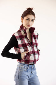 A woman wearing a burgundy, white, and black plaid zip-up fleece vest over a black long-sleeve top and light blue jeans, posing against a solid off-white background.