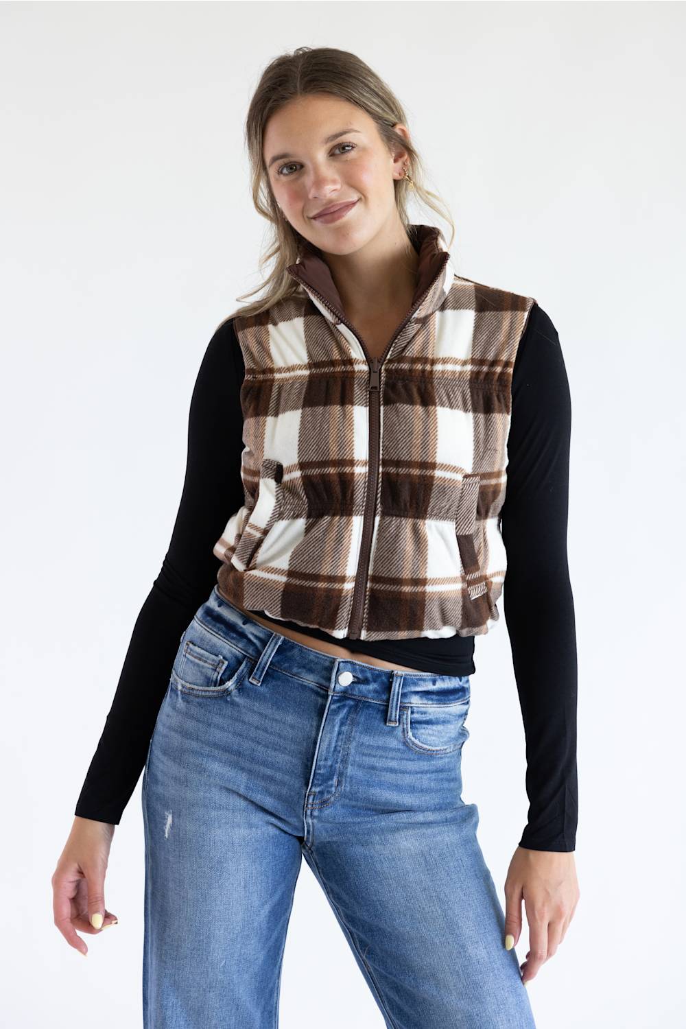 A woman modeling a brown and beige plaid zip-up fleece vest over a black long-sleeve shirt and blue jeans, photographed against a plain white studio background.