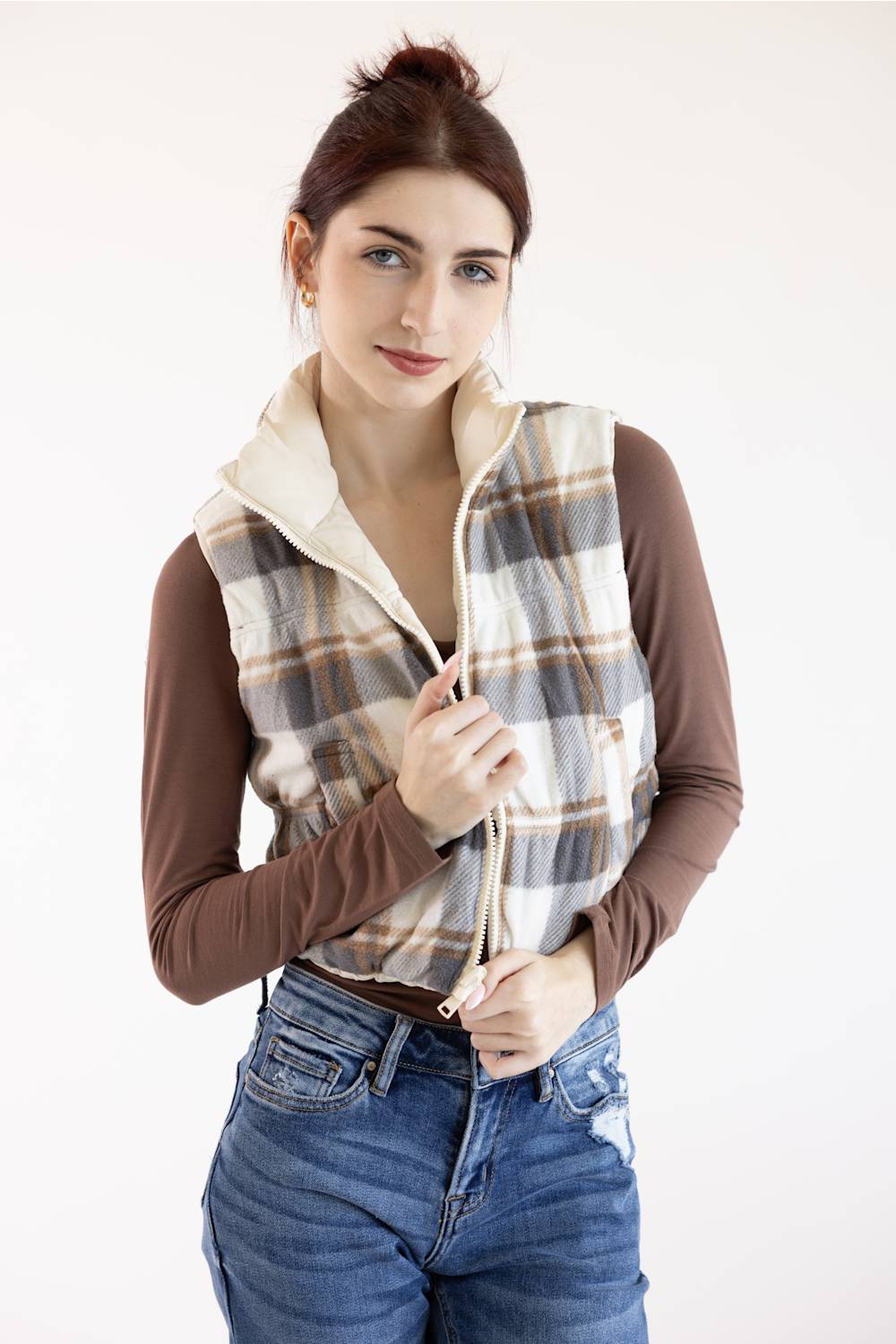 A woman modeling a sleeveless zip-front vest with a cream fleece collar and a beige, brown, and gray plaid pattern, worn over a brown long-sleeve top and blue jeans against a plain white background.