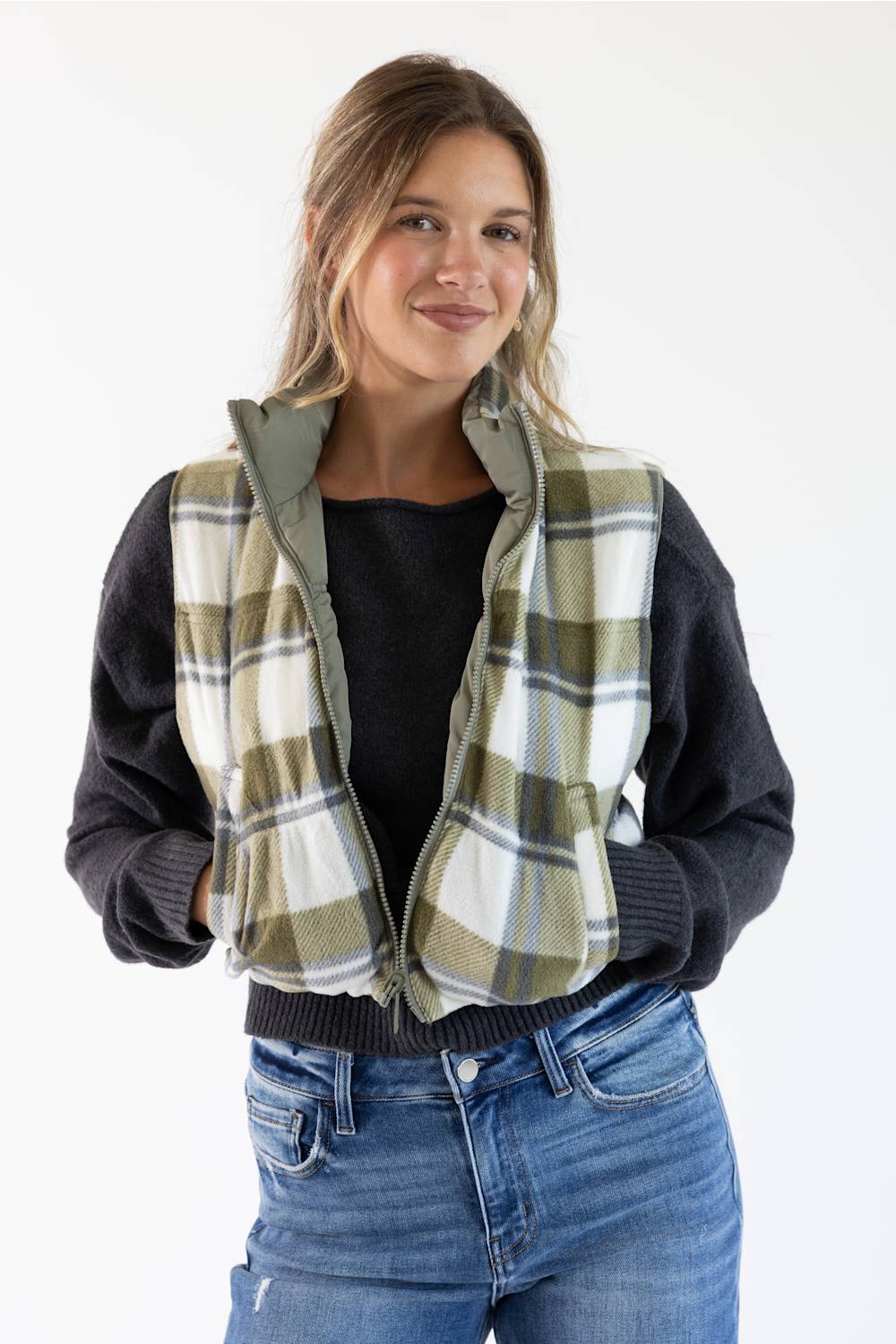 A woman wearing a zippered vest with a green, beige, and white plaid pattern over a black long-sleeve top and blue jeans, photographed against a plain white background.