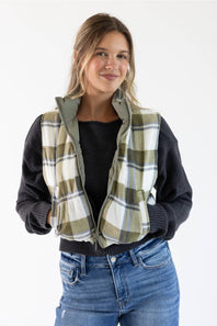 A woman wearing a zippered vest with a green, beige, and white plaid pattern over a black long-sleeve top and blue jeans, photographed against a plain white background.