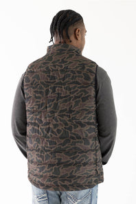 A man viewed from behind wearing a brown and black camouflage patterned quilted puffer vest over a dark gray long-sleeve shirt and blue jeans, against a plain white background.