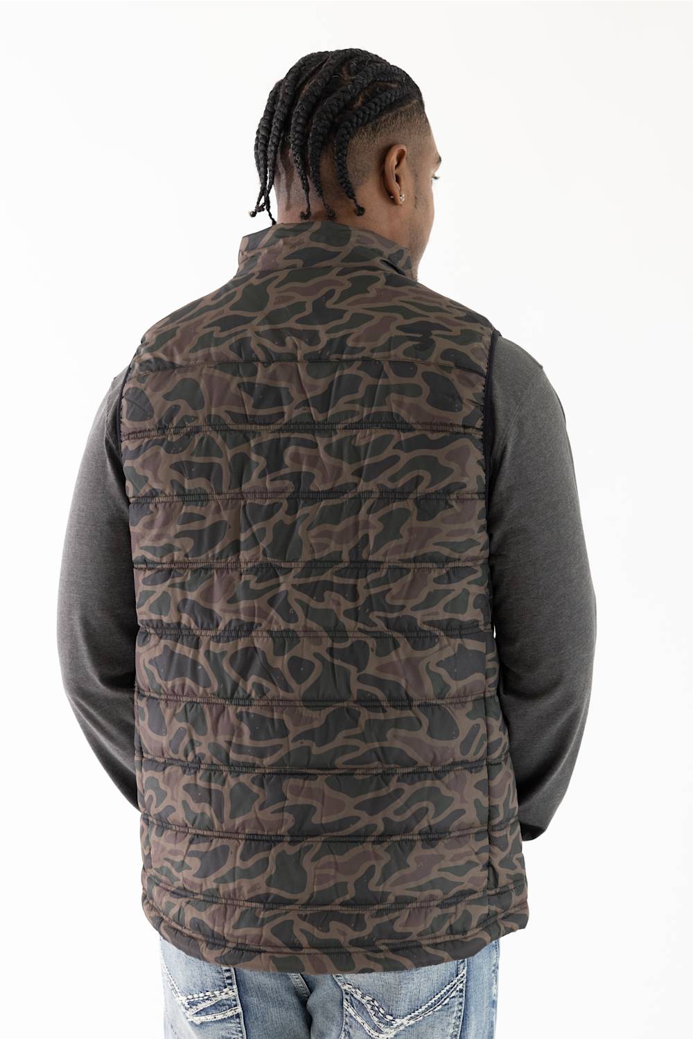 A man viewed from behind wearing a brown and black camouflage patterned quilted puffer vest over a dark gray long-sleeve shirt and blue jeans, against a plain white background.