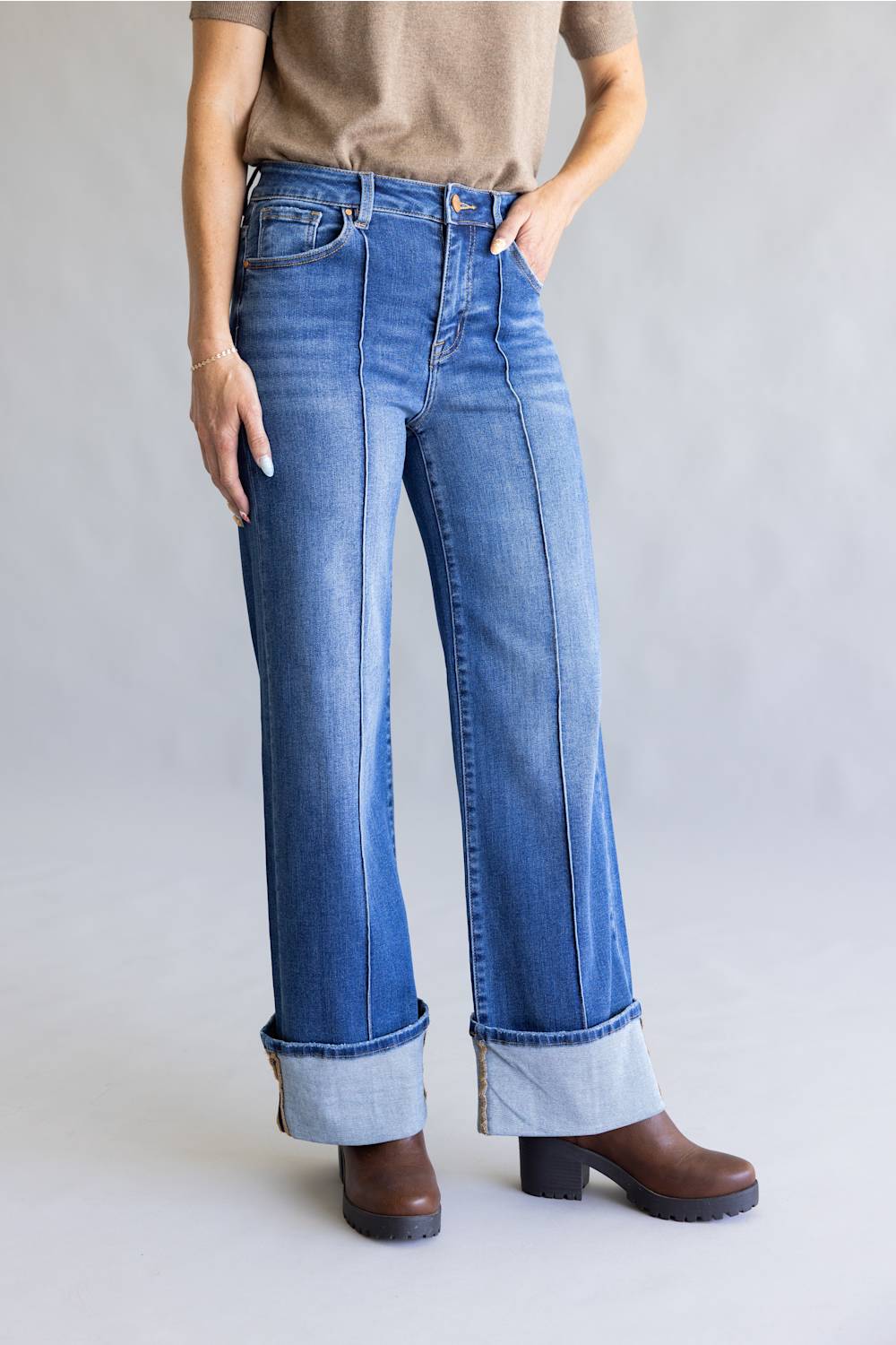 A person wearing blue wide-leg cuffed jeans with a tan top and brown boots, standing against a light gray studio background.