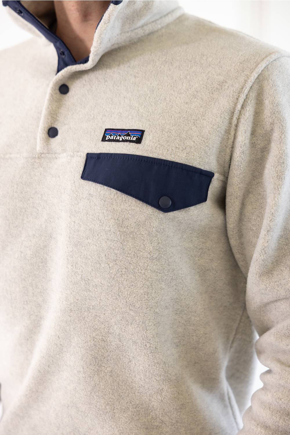 A man wearing a light beige fleece pullover sweater with a navy blue chest pocket and trim, paired with blue jeans, photographed against a plain white studio background.