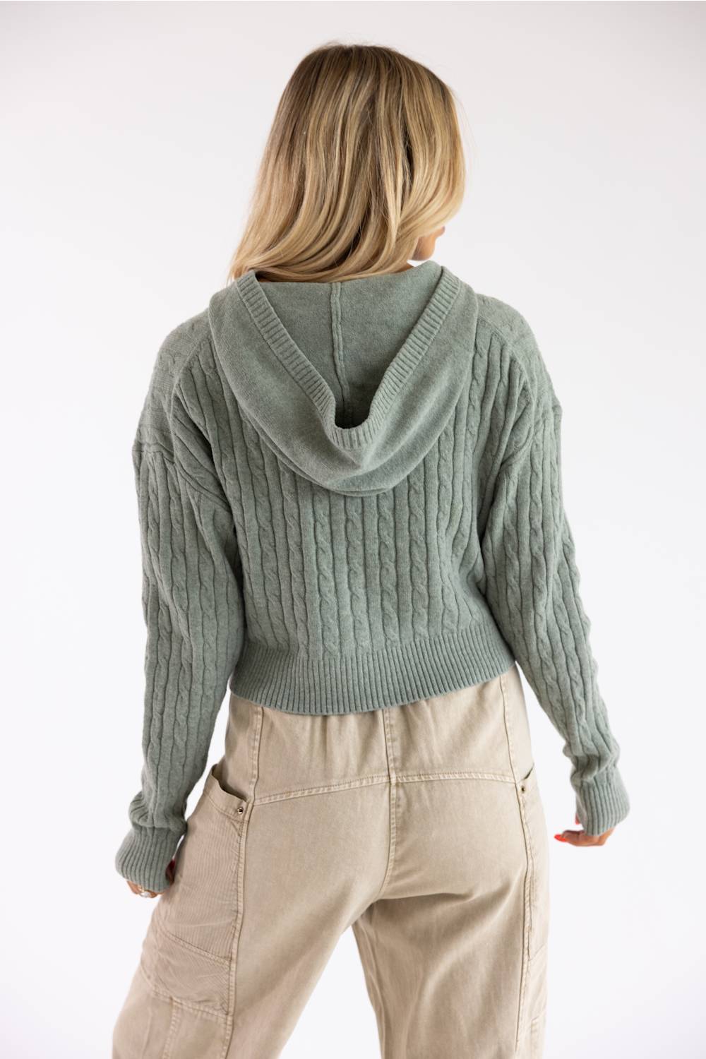 Close-up shot of a blonde woman wearing the Pink Rose cable knit hooded full-zip sweater in willow green, styled with beige drawstring pants.