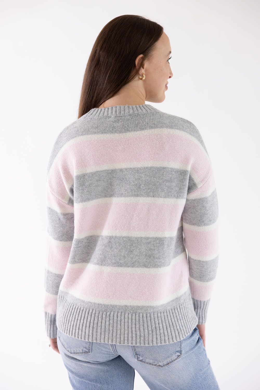 Back view of a woman wearing the Pink Rose Drop Shoulder Striped Knit Sweater in light pink, grey, and white horizontal stripes, featuring ribbed cuffs and hem, paired with light-wash jeans.