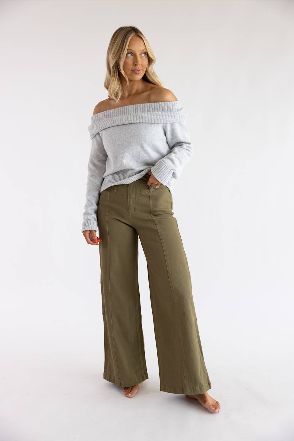 Full-length view of a blonde model standing barefoot in the Pink Rose heather grey off-the-shoulder knit sweater, styled with olive green high-waisted wide-leg trousers.