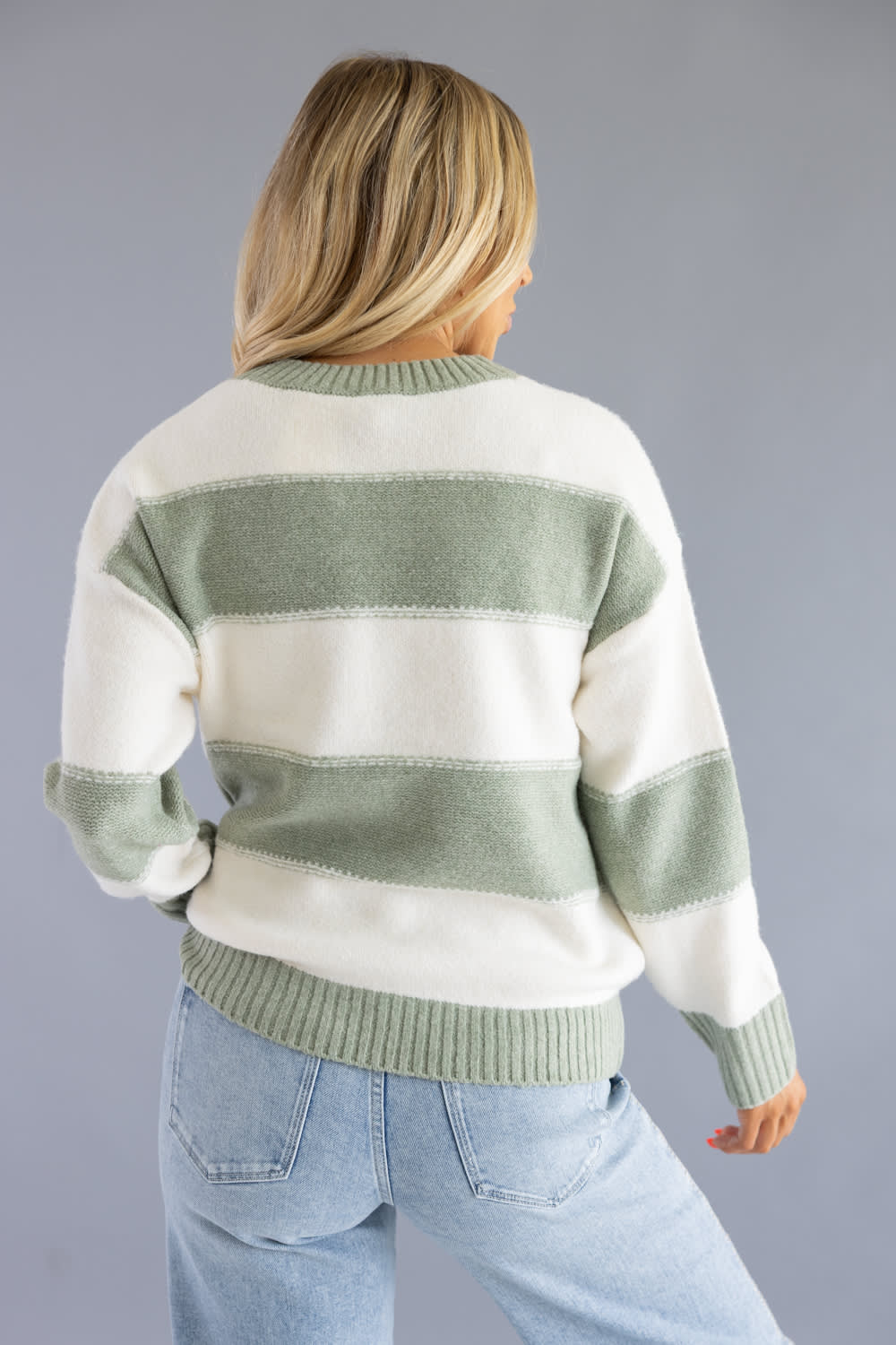Back view of a blonde woman wearing the Pink Rose cream and sage green horizontal striped crew neck sweater with light-wash jeans.