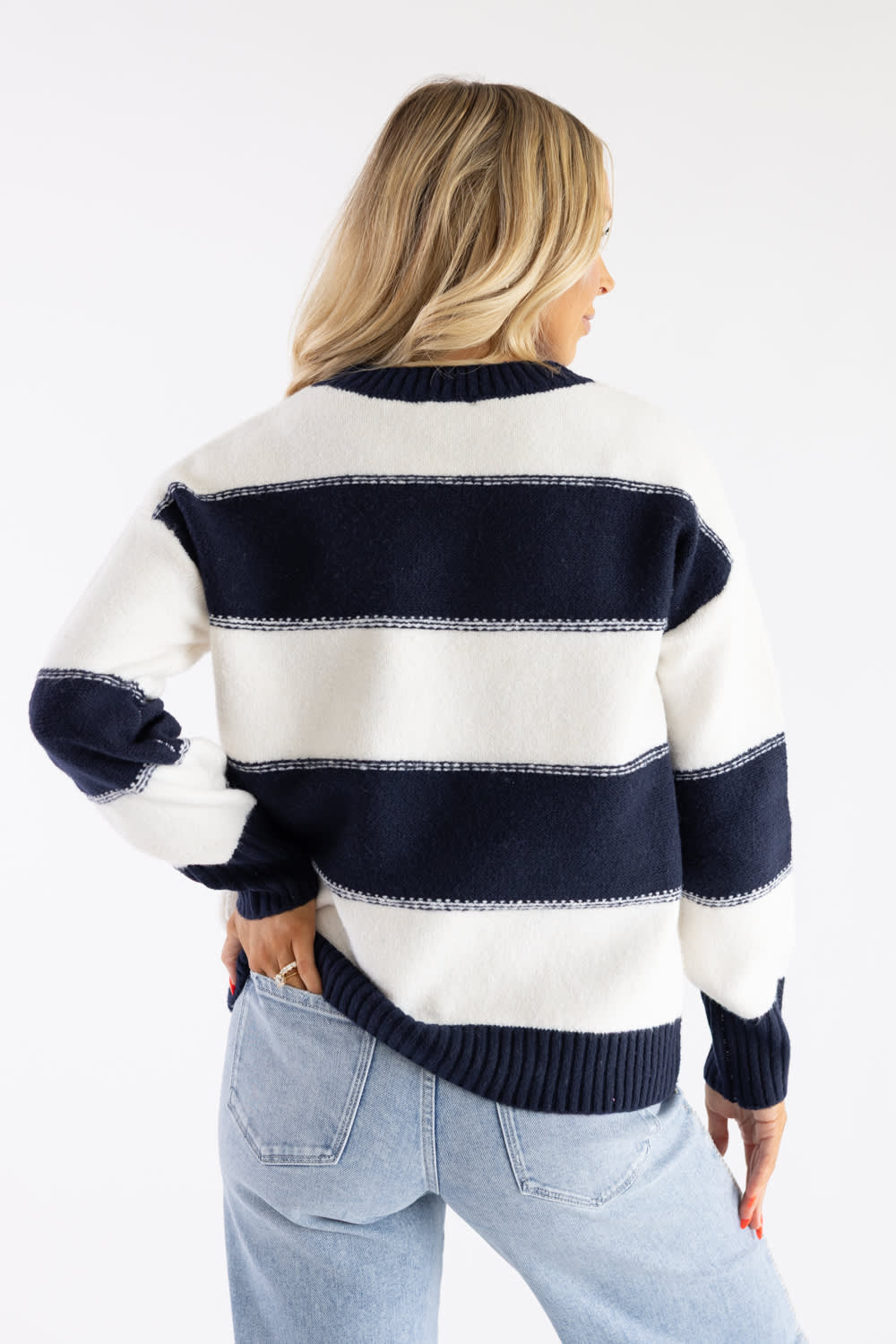 Back view of a blonde female model wearing the Pink Rose wide-striped crew neck knit sweater in cream and navy blue, showing the horizontal stripe pattern across the back and light wash denim jeans.