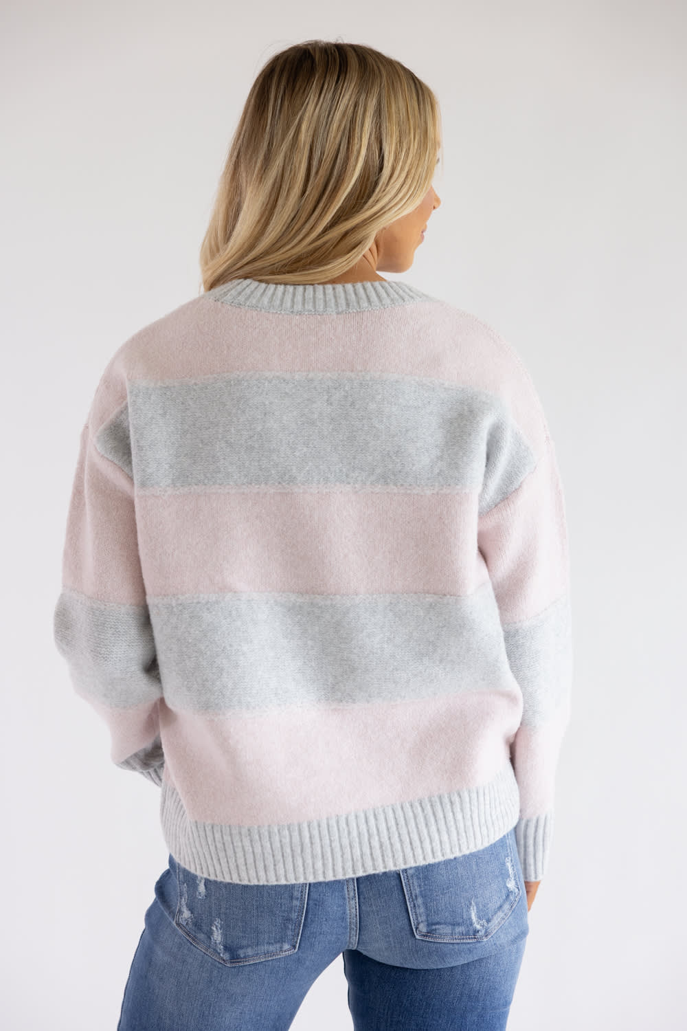 Back view of a blonde woman wearing the Pink Rose heather grey and light pink horizontal striped crew neck sweater with blue jeans.