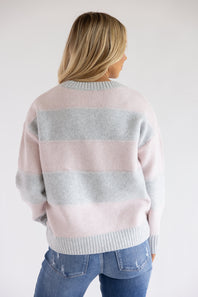 Back view of a blonde woman wearing the Pink Rose heather grey and light pink horizontal striped crew neck sweater with blue jeans.