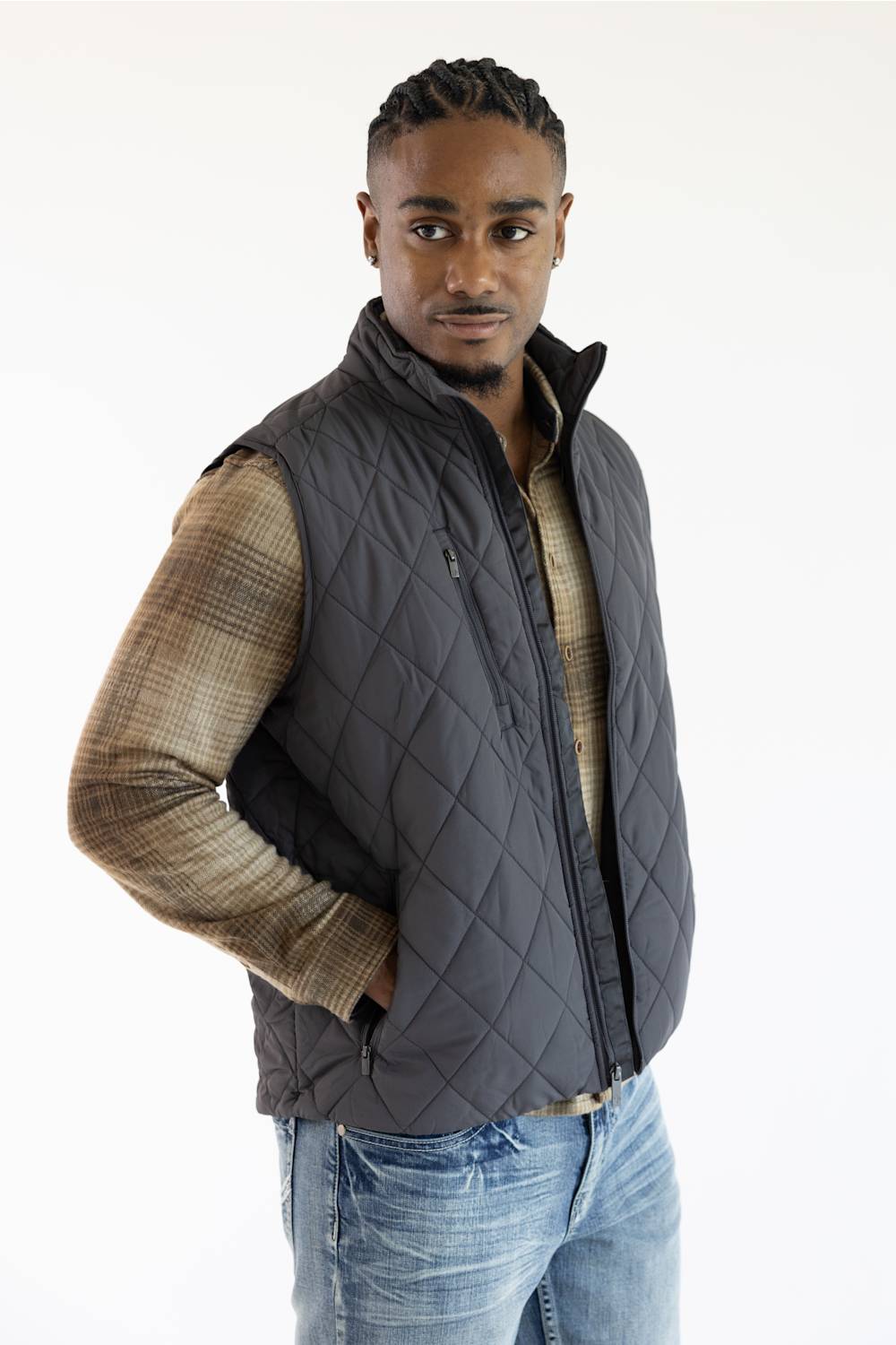 A man wearing a dark gray diamond-quilted zip-up puffer vest over a brown and beige plaid long-sleeve shirt and blue jeans, photographed against a plain white studio background.