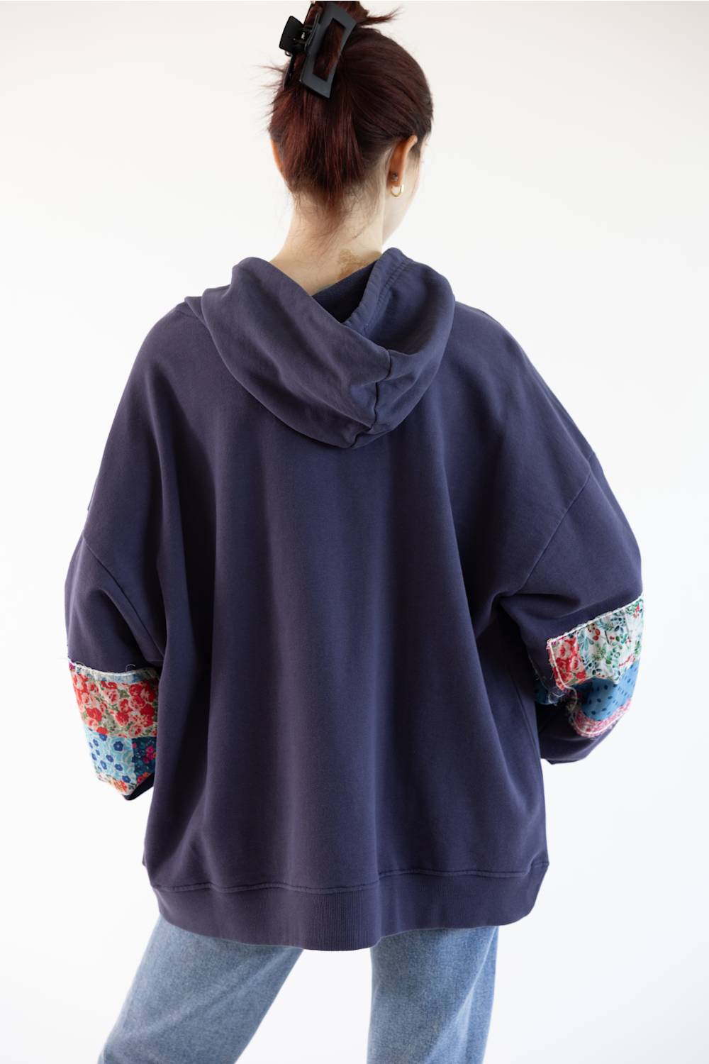 A person shown from the back wearing an oversized navy hoodie with colorful floral patchwork details on the sleeves, paired with light blue jeans against a plain white background.