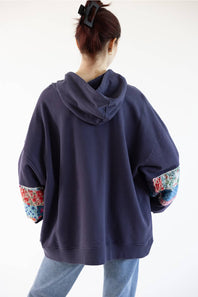 A person shown from the back wearing an oversized navy hoodie with colorful floral patchwork details on the sleeves, paired with light blue jeans against a plain white background.