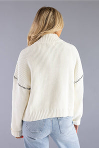 A woman viewed from the back wearing a cream ribbed knit mock-neck sweater with black dotted trim details on the sleeves, paired with light blue jeans, against a solid gray studio background.