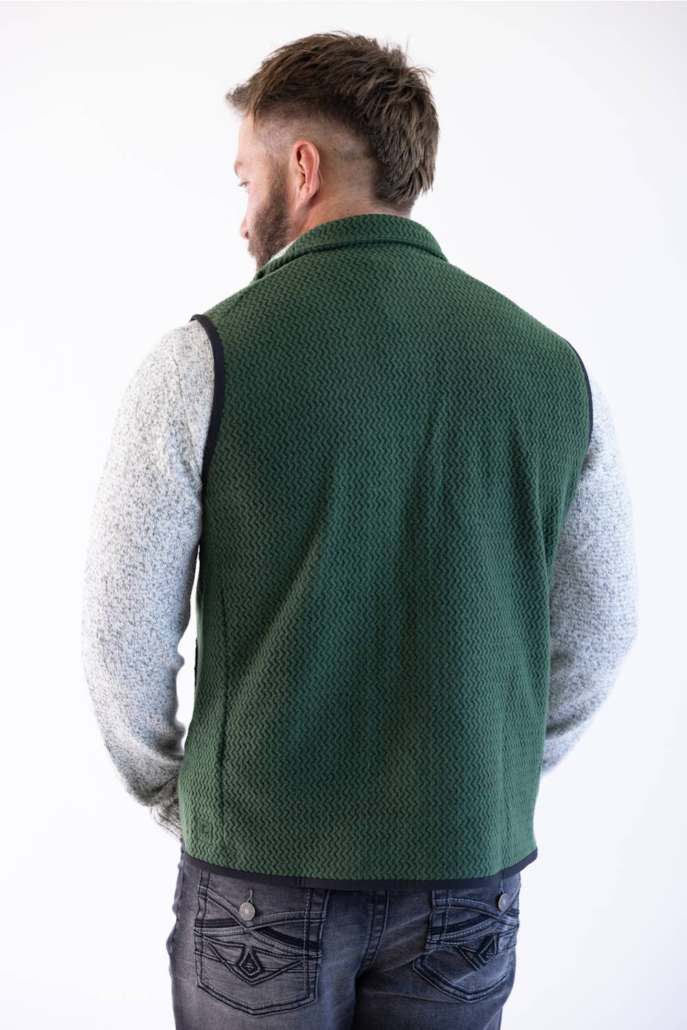 A man viewed from behind wearing a dark green quilted puffer vest over a beige long-sleeve shirt and blue jeans, against a plain white studio background.