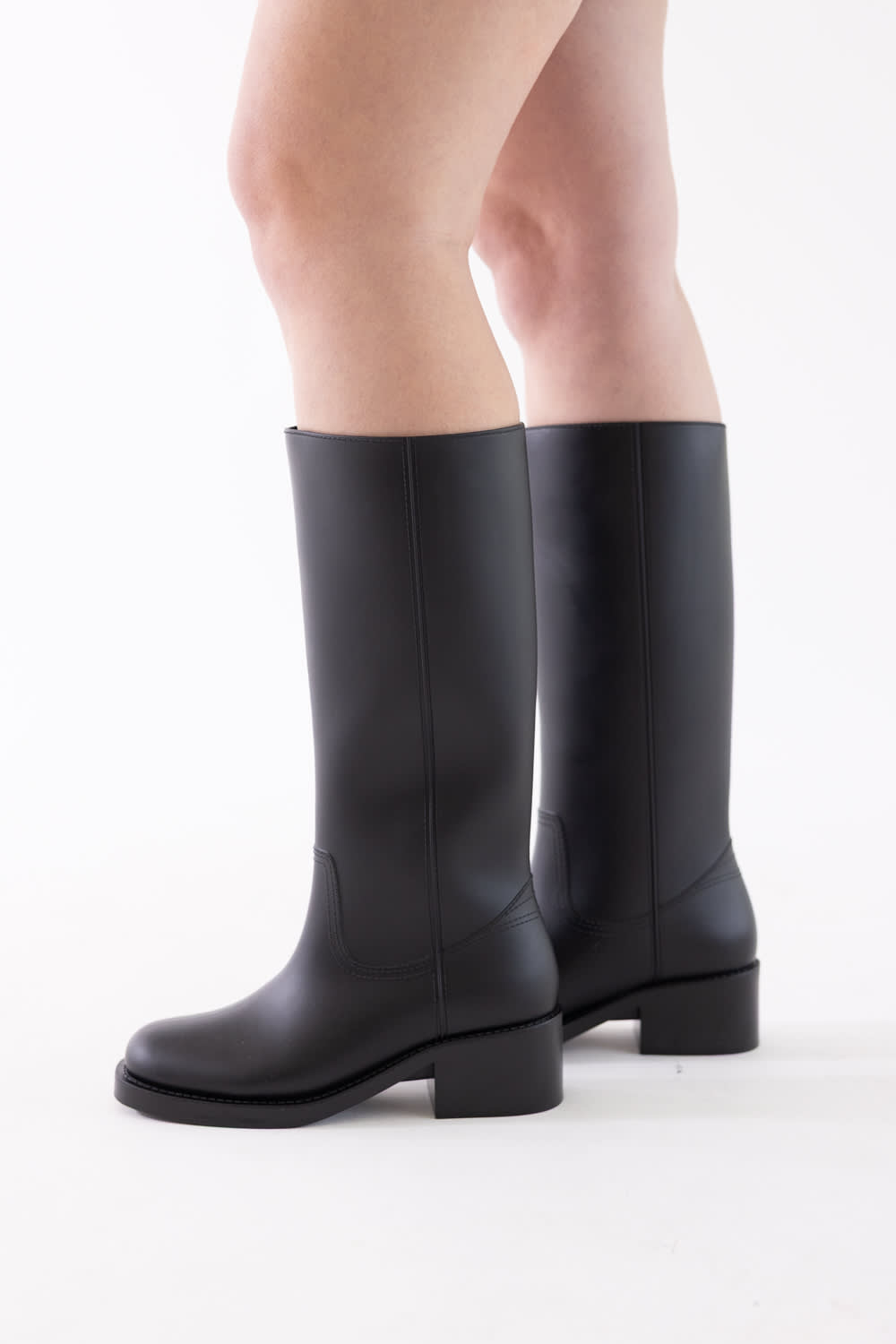 A pair of black leather knee-high boots with low block heels, shown on a model's feet against a plain white studio background.