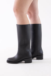 A pair of black leather knee-high boots with low block heels, shown on a model's feet against a plain white studio background.
