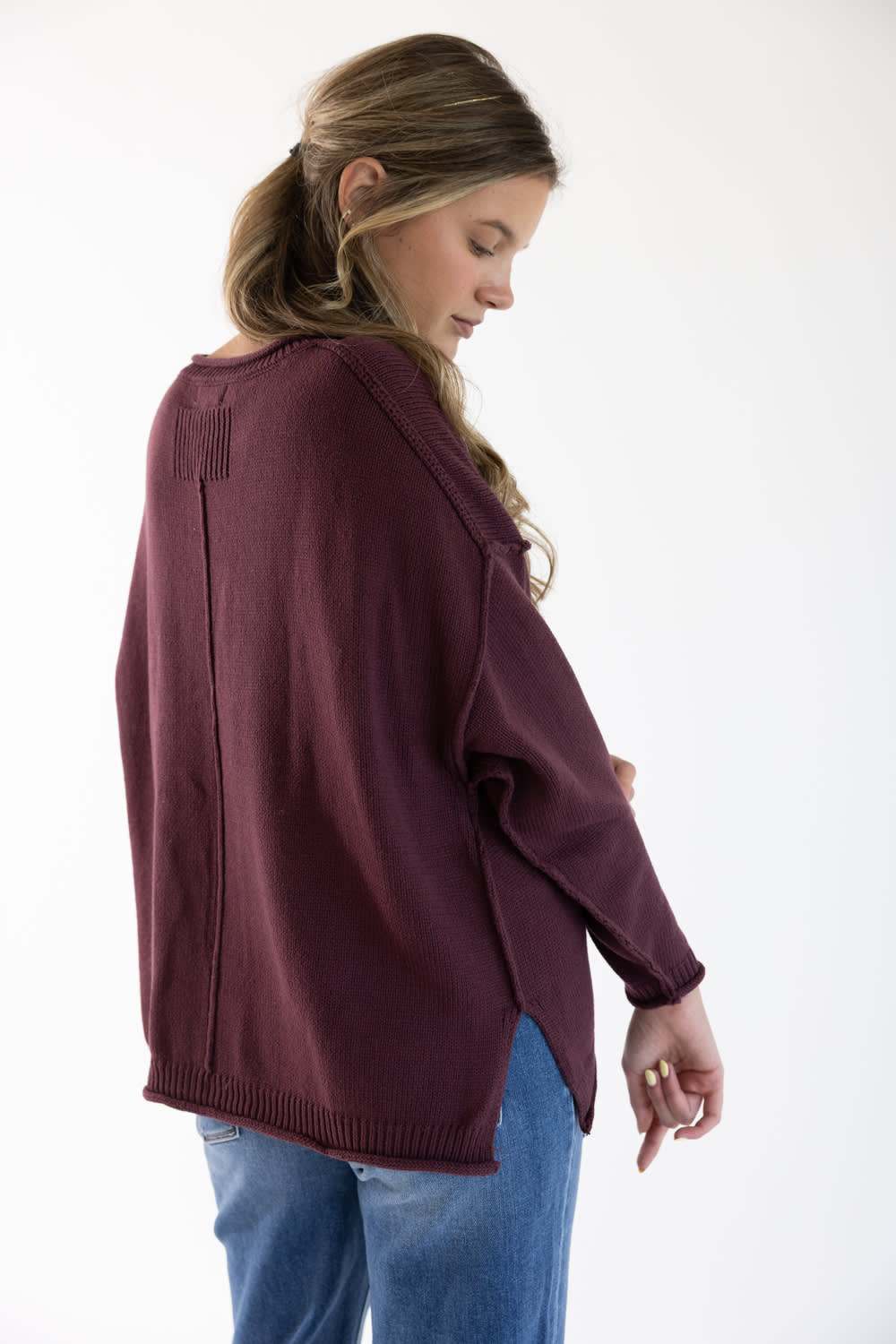 A woman viewed from the back wearing a maroon colored slouchy sweater with blue denim jeans standing in front of a white studio background