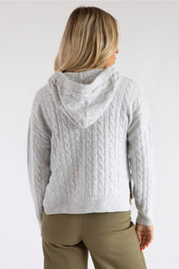 A woman viewed from the back wearing a light gray cable-knit hooded sweater and olive green pants, set against a plain light gray studio background.