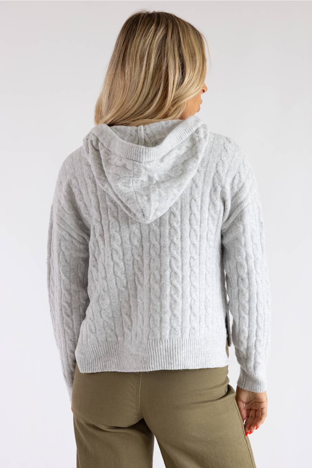 A woman viewed from the back wearing a light gray cable-knit hooded sweater and olive green pants, set against a plain light gray studio background.