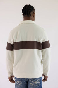 A man viewed from behind wearing a cream-colored long-sleeve rugby-style shirt with a wide dark brown horizontal stripe across the upper arms, paired with blue jeans.