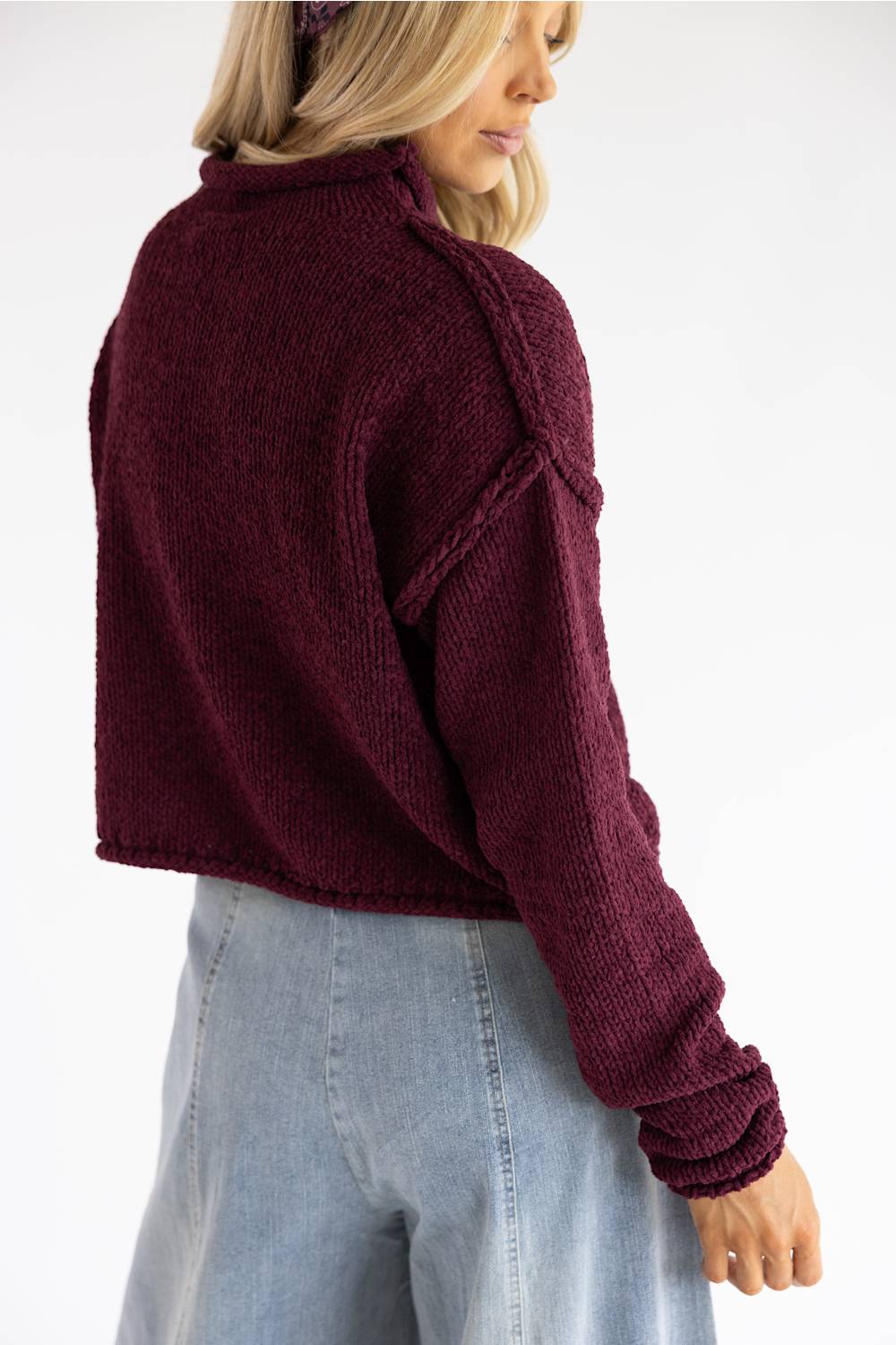 A woman wearing a cropped, long-sleeve knit sweater in deep burgundy, shown from a side-back angle against a plain white background.