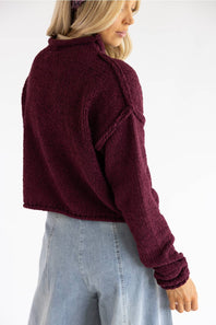 A woman wearing a cropped, long-sleeve knit sweater in deep burgundy, shown from a side-back angle against a plain white background.