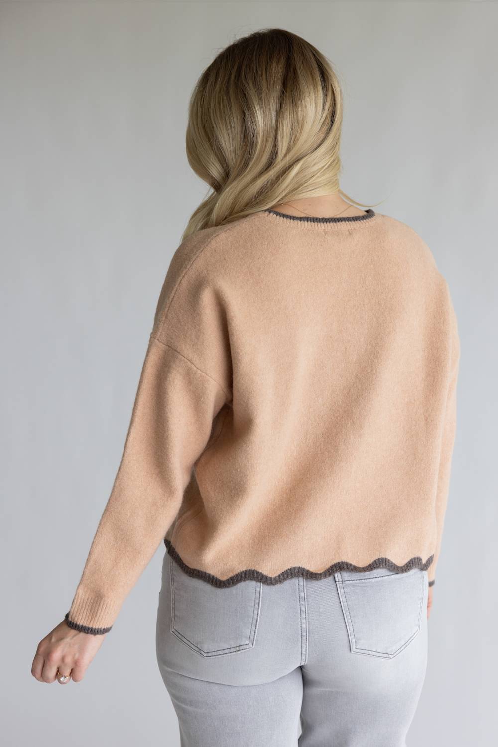 A woman viewed from the back wearing a light peach-beige knit sweater with dark gray scalloped trim at the neckline and hem, paired with light gray jeans, against a plain light gray background.