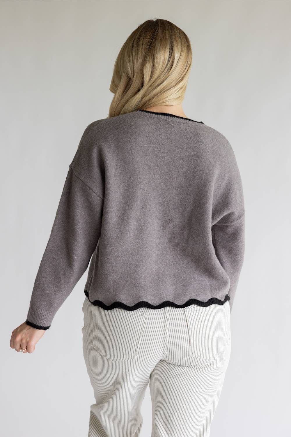 A woman shown from the back wearing a taupe gray knit sweater with black scalloped trim and white ribbed pants, photographed against a plain light gray studio background.