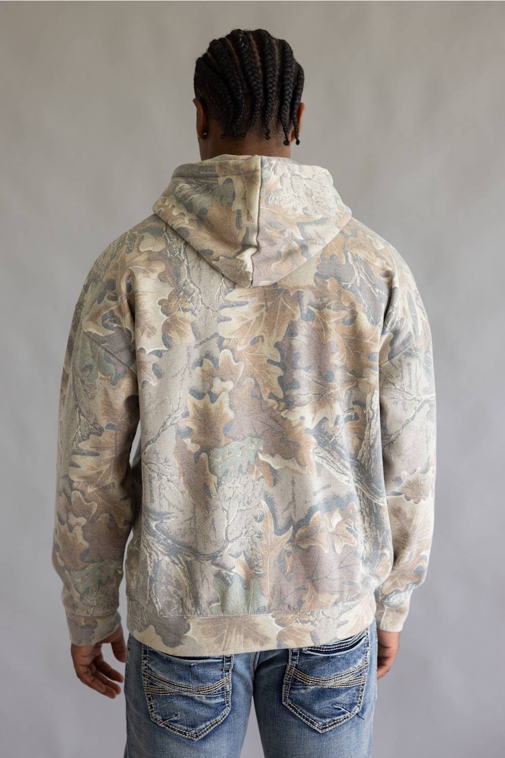A man viewed from behind wearing a hooded sweatshirt with an abstract leaf-like camouflage pattern in beige, gray, and muted blue-green tones, paired with blue jeans against a plain light gray studio background.