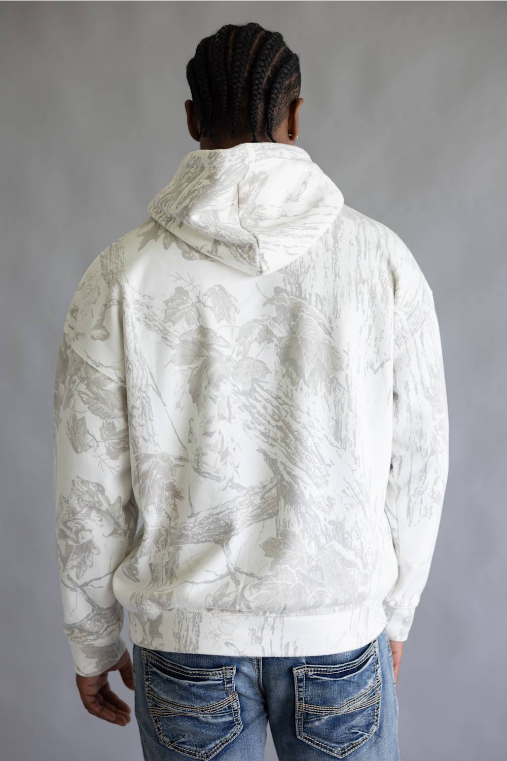 A man viewed from behind wearing a white hooded sweatshirt with a light gray camo pattern, paired with blue jeans, against a neutral gray studio background.