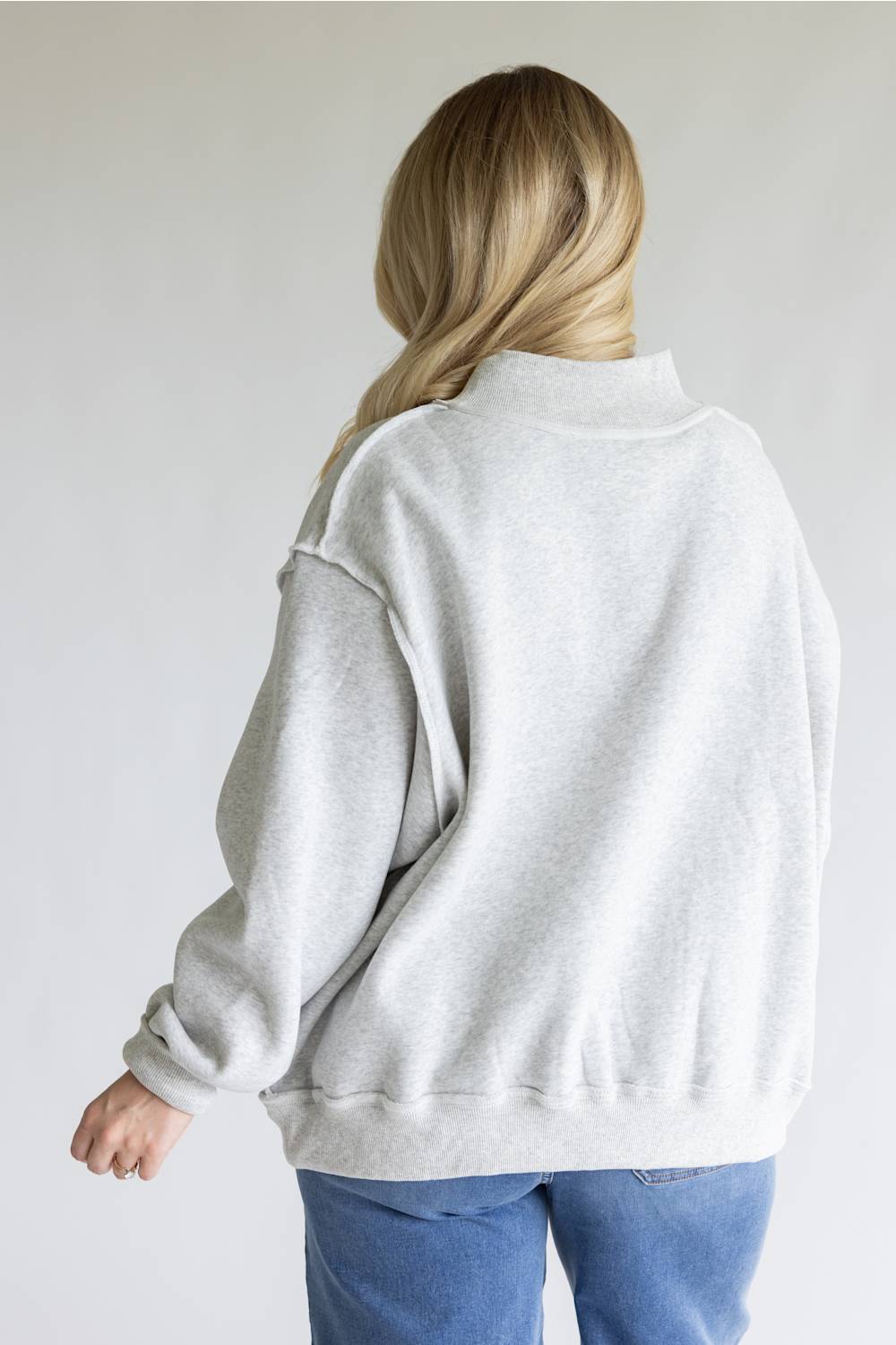 A woman viewed from behind wearing an oversized light gray heather sweatshirt and blue jeans against a plain light gray background.