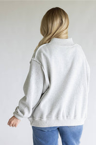 A woman viewed from behind wearing an oversized light gray heather sweatshirt and blue jeans against a plain light gray background.